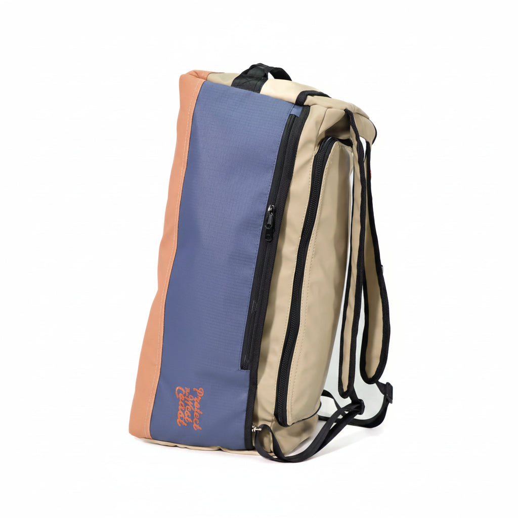 A color-blocked backpack with a slate blue front panel, beige side panels, and peach-colored side accents, featuring black zippers and straps, shown on a white background.