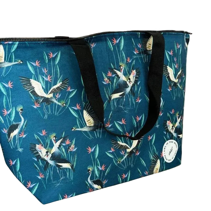 A large tote bag with a dark teal blue background, featuring a pattern of flying cranes, green reeds, and pink flowers. The bag has black webbed shoulder straps and a black zipper closure,